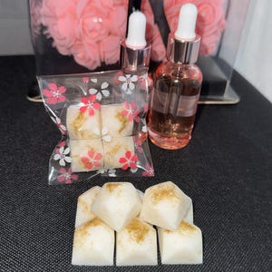 Peut inclure: Six cubes de cire blanche avec des paillettes dorées, emballés dans un sac en plastique transparent avec un motif floral. Deux flacons en verre avec des compte-gouttes, l'un avec un liquide rose et l'autre avec un liquide clair, sont à l'arrière-plan.