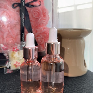 Peut inclure: Deux flacons en verre transparent contenant un liquide rose et des compte-gouttes argentés et blancs. Les flacons sont posés sur une surface noire.