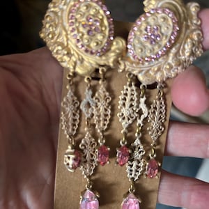 May include: Ornate gold-tone earrings with pink accents. The earrings feature a detailed filigree design with dangling pink teardrop-shaped gems. The top portion has a decorative oval with pink crystals. A vintage-inspired accessory.