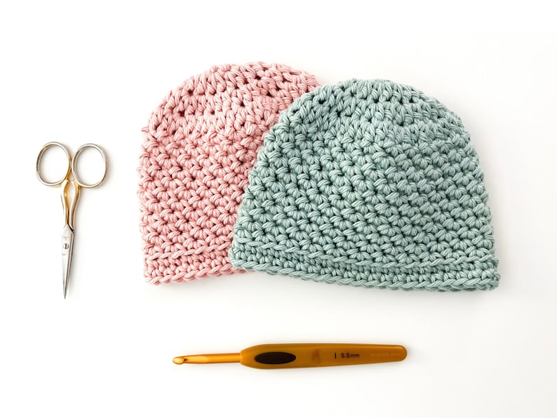 May include: Two crocheted hats, one pink and one light blue, with a gold crochet hook and a pair of silver scissors on a white background.
