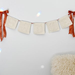 May include: A white crocheted banner with five squares, each with a textured pattern, hangs on a white wall. The banner is decorated with two orange satin bows on either end.