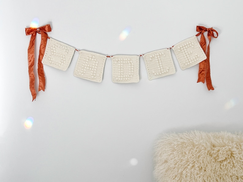 May include: A white crocheted banner with five squares, each with a textured pattern, hangs on a white wall. The banner is decorated with two orange satin bows on either end.
