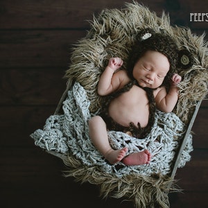 May include: A newborn baby wearing a brown crocheted bear hat and matching outfit is sleeping in a basket lined with a white crocheted blanket and brown faux fur.