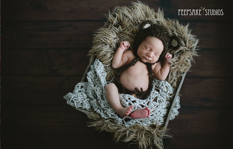 May include: A newborn baby wearing a brown crocheted bear hat and matching outfit is sleeping in a basket lined with a white crocheted blanket and brown faux fur.
