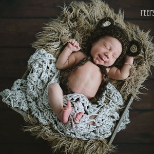 May include: A newborn baby wearing a brown crocheted bear hat with floppy ears, lying on a white crocheted blanket in a brown faux fur-lined basket. The baby is smiling and has its arms outstretched.