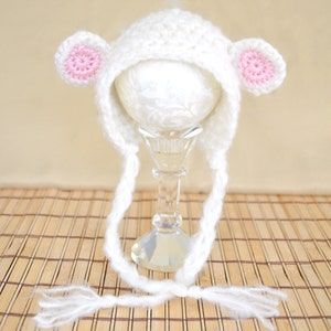 May include: White crocheted baby hat with pink ears and long ties. The hat is on a stand and has a white background.