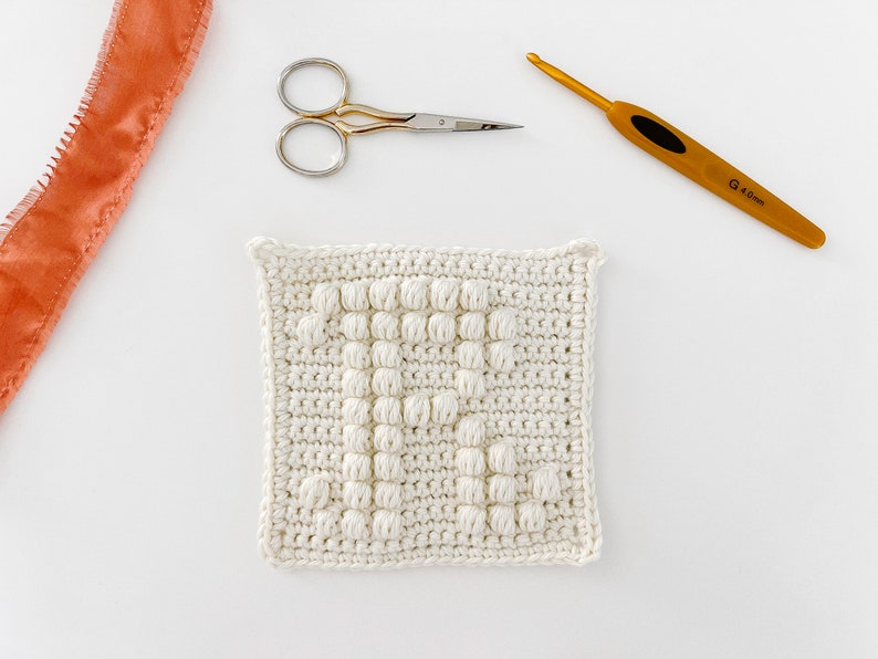 May include: A white crocheted square with a textured pattern, a pair of silver scissors, and a gold crochet hook labeled "4.0mm".
