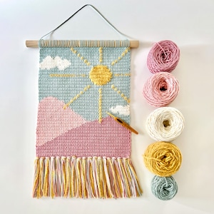 May include: A crocheted wall hanging with a sun, clouds, and mountains design. The hanging is made with pink, blue, white, and yellow yarn. There are five balls of yarn in the colors used in the hanging.