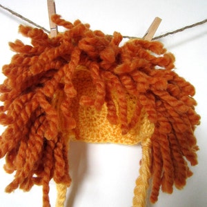 May include: A crocheted lion hat with a yellow base and orange yarn mane. The hat is hanging on a clothesline.