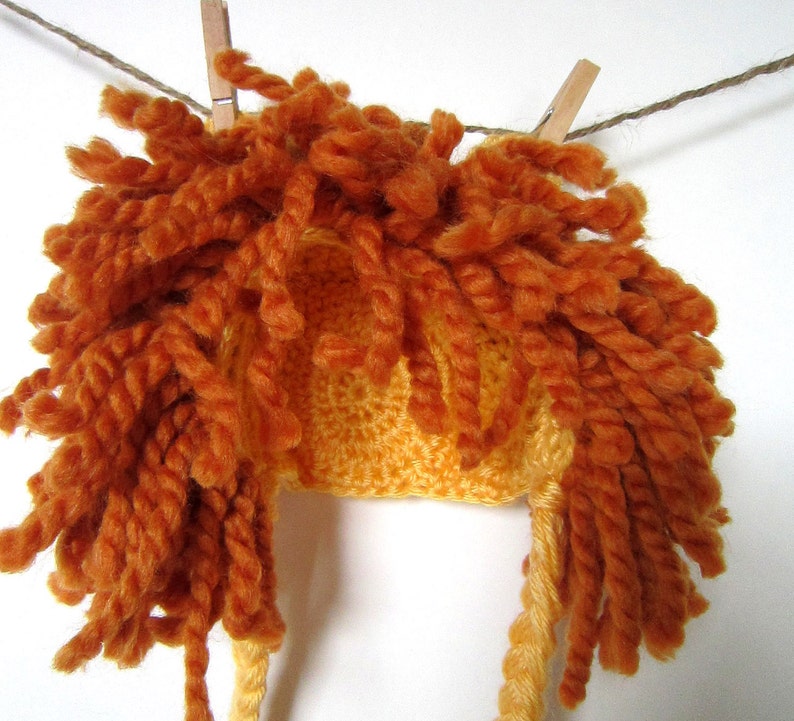 May include: A crocheted lion hat with a yellow base and orange yarn mane. The hat is hanging on a clothesline.