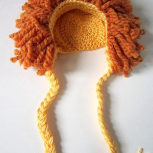 May include: A crocheted lion hat with a yellow mane and orange fur. The hat has two long yellow braids with tassels at the ends.