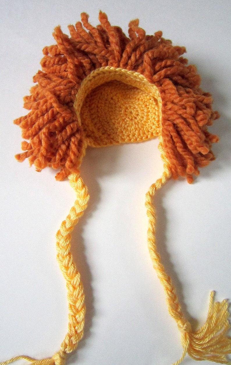 May include: A crocheted lion hat with a yellow mane and orange fur. The hat has two long yellow braids with tassels at the ends.