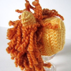 May include: A crocheted hat with a yellow body and orange yarn mane. The hat is on a clear glass stand.
