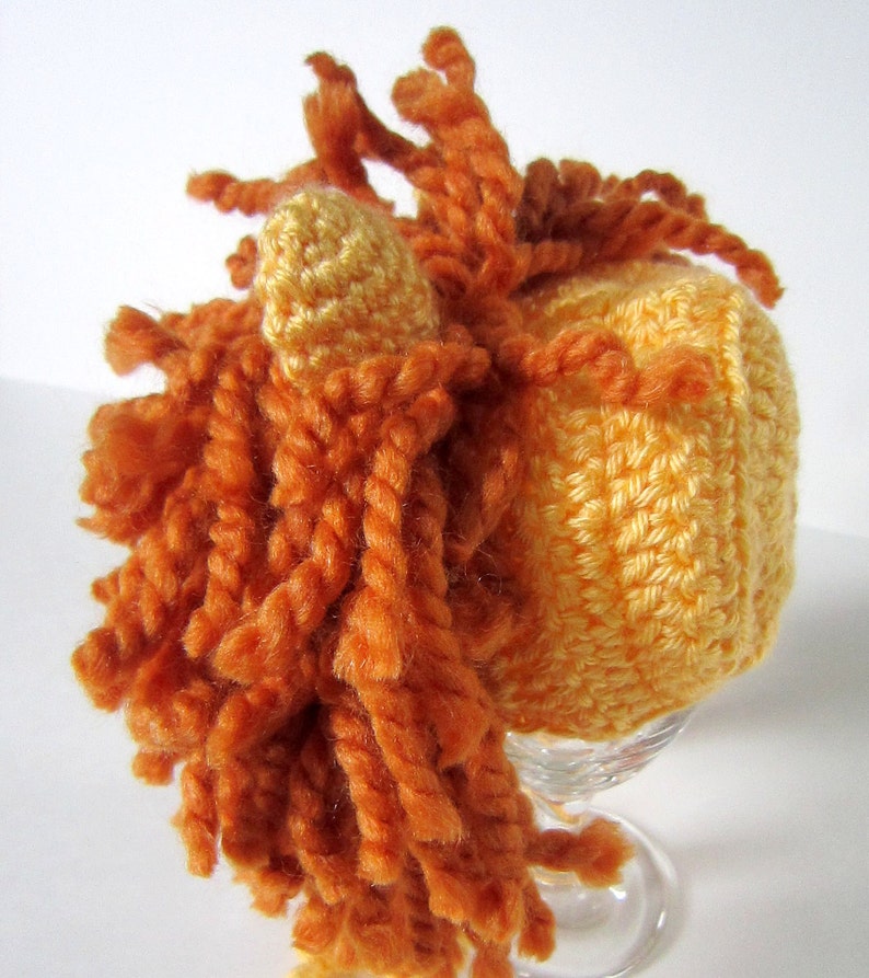 May include: A crocheted hat with a yellow body and orange yarn mane. The hat is on a clear glass stand.