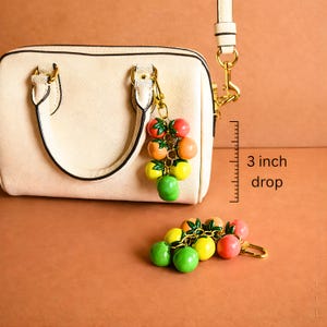 Puede incluir: Un bolso crema con cierre y asa dorados, acompa&ntilde;ado de un charm de frutas. El charm presenta un racimo de frutas rojas, naranjas, amarillas y verdes. La longitud de la ca&iacute;da del charm es de 7,6 cm.