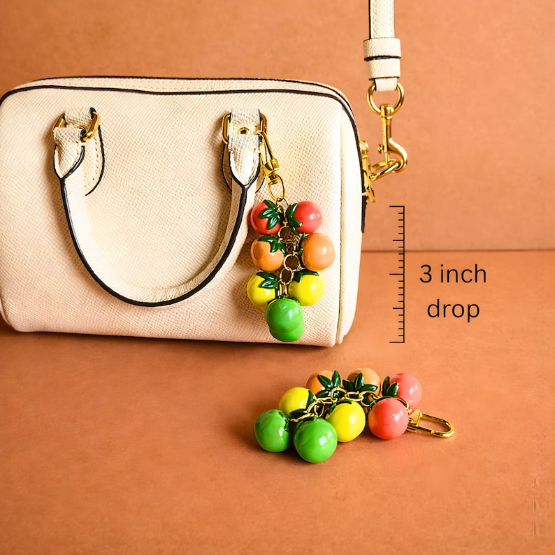 Puede incluir: Un bolso crema con cierre y asa dorados, acompa&ntilde;ado de un charm de frutas. El charm presenta un racimo de frutas rojas, naranjas, amarillas y verdes. La longitud de la ca&iacute;da del charm es de 7,6 cm.