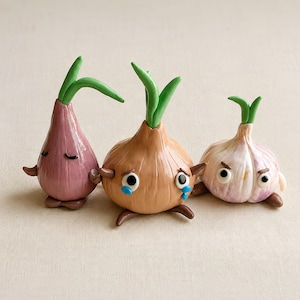 Pode incluir: Três pequenas figuras de vegetais antropomórficos com brotos verdes no topo. À esquerda, uma cebola roxa alongada com olhos fechados. Ao centro, uma cebola bege redonda com lágrimas azuis. À direita, um bulbo de alho branco.