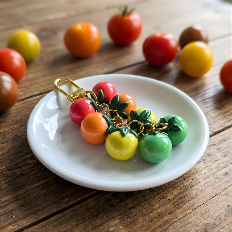 Puede incluir: Un llavero colorido con un grupo de dijes de tomate en miniatura en rojo, naranja, amarillo y verde. Los dijes tienen detalles de hojas verdes y est&aacute;n unidos a un cierre dorado. El llavero est&aacute; sobre un plato blanco, con tomates dispersos en el fondo.