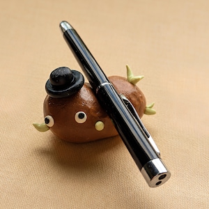 May include: A brown, potato-shaped clay figure with a black bowler hat, two white eyes with black pupils, and small light green protrusions, serving as a holder for a sleek black and silver ballpoint pen resting diagonally across its body.