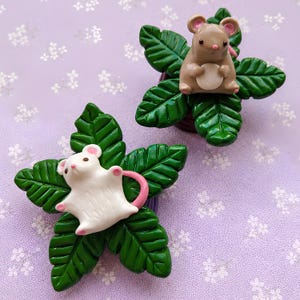 May include: Two decorative ornaments featuring small sculpted mice resting on dark green leaves. One mouse is light brown with a cream belly, and the other is white with a pink tail. Both figures have small black eyes and pink accents on their ears.