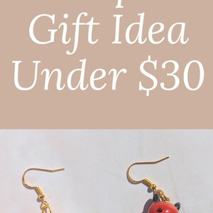 Puede incluir: Pendientes hechos a mano con un diablo rojo y un &aacute;ngel blanco, ambos con ganchos dorados. Los pendientes se muestran en una tarjeta blanca con el texto "Handmade with love".