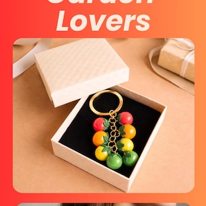 Puede incluir: Un llavero colorido con dijes de frutas rojas, naranjas, amarillas y verdes. Tiene un cierre y una cadena dorados. Tambi&eacute;n se muestra el texto "Cute Summer Accessory Gift for Garden Lovers".
