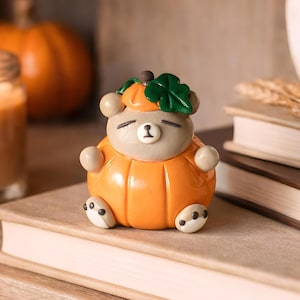 May include: A charming bear figurine in an orange pumpkin costume, with a green leaf and brown stem. The bear has a light brown body, a white face, and black eyes. It is resting on a book. The figurine is a cute autumn decoration.