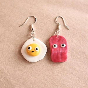 May include: A pair of novelty earrings, one shaped like a fried egg and the other like a piece of bacon. The egg has a yellow yolk and white egg white. Both have black and white eyes and silver-coloured ear wires.