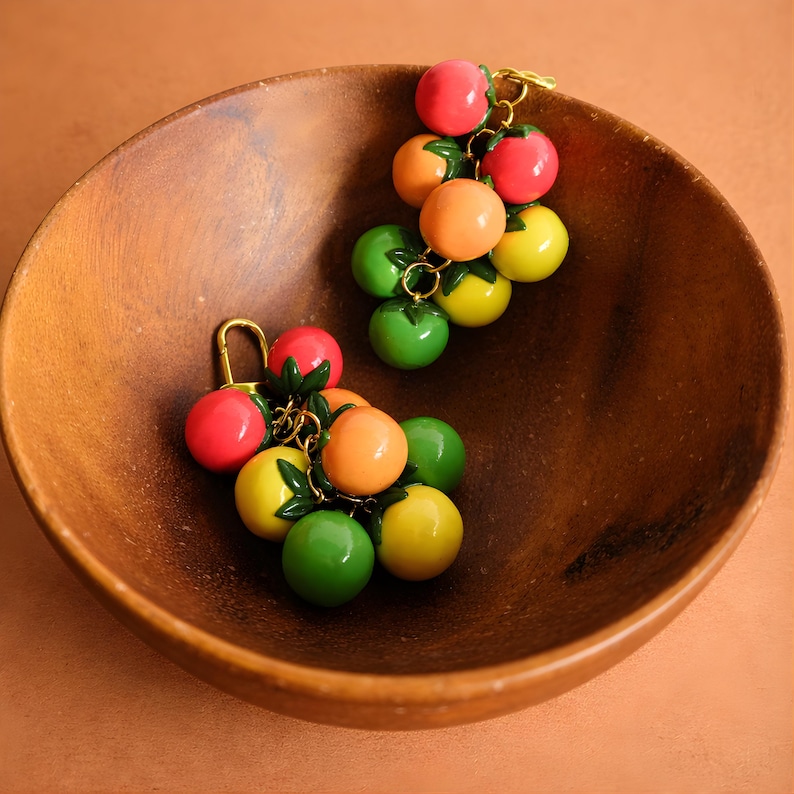 Puede incluir: Dos conjuntos de colgantes coloridos con forma de fruta, que parecen uvas, descansan en un cuenco de madera. Cada colgante presenta un grupo de esferas vibrantes y brillantes en rojo, naranja, verde y amarillo, conectadas por metal dorado. El cuenco es marr&oacute;n.