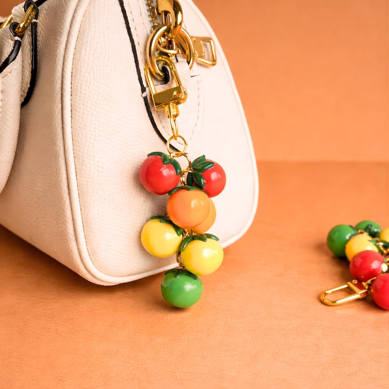 Puede incluir: Un colorido charm con un racimo de tomates rojos, naranjas, amarillos y verdes, sujeto a un cierre dorado. El charm cuelga de un bolso crema con herrajes dorados, sobre un fondo naranja.