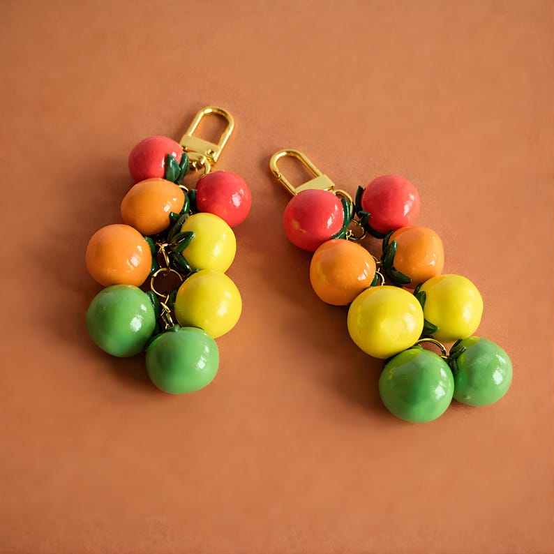 Puede incluir: Dos llaveros con racimos de colgantes de frutas redondas y coloridas. Cada colgante es de un color diferente, incluyendo rojo, naranja, amarillo y verde. Los llaveros tienen cierres dorados y peque&ntilde;as hojas verdes.