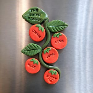 May include: A personalized family name magnet in the shape of a tomato plant. The magnet features orange tomatoes with individual names and green leaves. The top leaf reads "THE SMITH FAMILY".