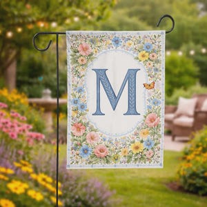 May include: A garden flag featuring a large blue letter "M" in an oval frame, surrounded by a floral border of pink, blue, and yellow flowers. A small orange butterfly is visible. The flag is displayed on a black metal stand.