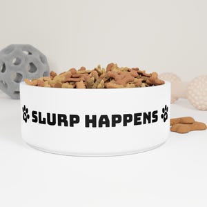 以下が含まれることがあります： 犬用のドライフードが入った白いセラミック製の犬用ボウル。「SLURP HAPPENS」というフレーズが黒い文字で書かれており、両側に足跡が付いています。他の犬のおもちゃが背景にあります。