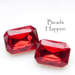 May include: Two faceted, rectangular, bright red gemstones. The gems are displayed on a white background. The text "Beads Happen" is written in black script above the gemstones.