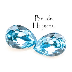 May include: Two pear-shaped, faceted, light blue gemstones are displayed against a white background. The gemstones are positioned close together, with the text "Beads Happen" above them.