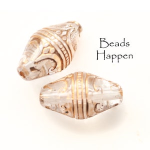May include: Two clear glass beads with gold accents and intricate designs. The beads are oval shaped and have a glossy finish. "Beads Happen" is written in black text on a white background.