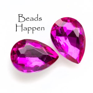Puede incluir: Dos cuentas de cristal de color rosa brillante en forma de l&aacute;grima. Las cuentas son facetadas y tienen un acabado brillante. El texto "Beads Happen" est&aacute; escrito en negro sobre las cuentas.