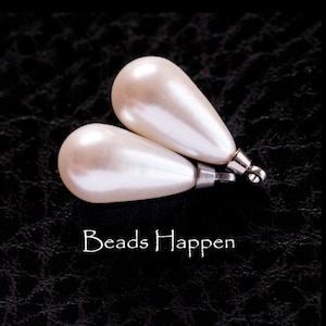 May include: Two pearlescent white teardrop-shaped beads with silver caps and loops. The beads are on a black textured surface. The text "Beads Happen" is written below the beads.
