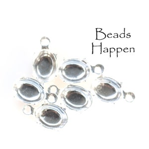 Könnte beinhalten: Silberne ovale Perlenkappen mit einer Schlaufe an jeder Seite. Die Perlenkappen sind in einer Gruppe auf einem weißen Hintergrund angeordnet. Der Text "Beads Happen" ist in Schwarz über den Perlenkappen gedruckt.
