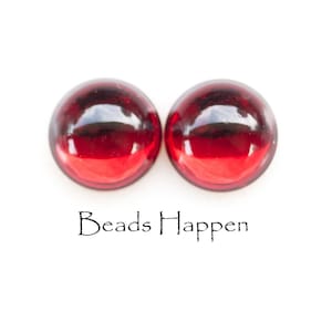 May include: Two round, domed, deep red glass beads. The beads are smooth and glossy, reflecting light. The text "Beads Happen" is printed in black script below the beads.