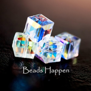 May include: Four clear, iridescent cube beads are clustered together, reflecting a spectrum of colors. The beads are faceted, catching and refracting light. The text "Beads Happen" is written in white script at the bottom.