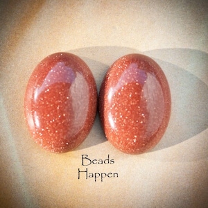 May include: Two oval-shaped, red-brown sandstone cabochons with a glittery finish. The cabochons are smooth and polished. The text "Beads Happen" is written below the cabochons.