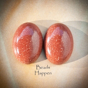 May include: Two oval-shaped, red-brown sandstone cabochons with a glittery finish. The cabochons are smooth and polished. The text "Beads Happen" is written below the cabochons.