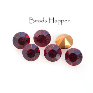 May include: Six red glass rhinestones and one gold rhinestone. The rhinestones are round and have a faceted design. The rhinestones are arranged in a row on a white background. The text "Beads Happen" is printed above the rhinestones.