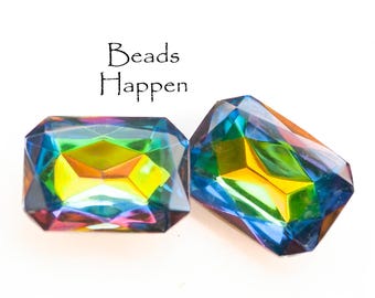 Puede incluir: Dos cuentas de vidrio facetadas iridiscentes en forma rectangular con un arcoíris de colores, incluyendo azul, verde, amarillo y naranja. El texto "Beads Happen" está escrito sobre las cuentas.