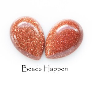 May include: Two teardrop-shaped, reddish-brown gemstone beads with a sparkly, glittery texture. The beads are arranged in a heart shape. The text "Beads Happen" is written below the beads.