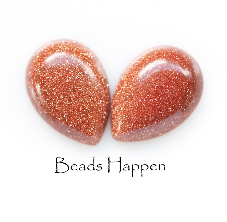 May include: Two teardrop-shaped, reddish-brown gemstone beads with a sparkly, glittery texture. The beads are arranged in a heart shape. The text "Beads Happen" is written below the beads.
