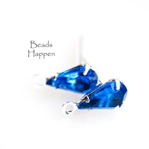 May include: Two teardrop-shaped blue crystal beads with silver settings and loops. The beads are on a white background.