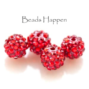 May include: Four red rhinestone beads, each with a round shape and a textured surface. The beads are arranged in a row, with the text "Beads Happen" above them.